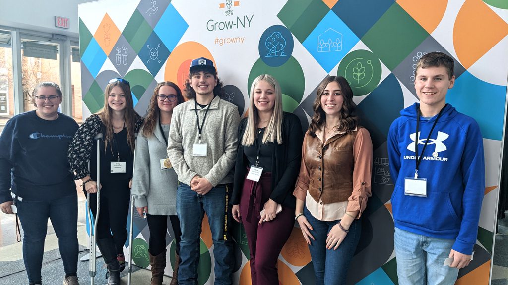 Seven students stand against a colorful backdrop decorated with the words Grow NY. 
