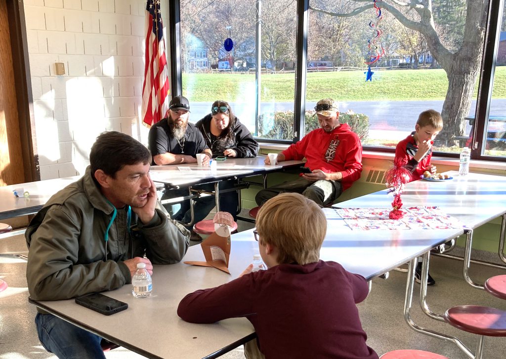 Adults and students sit and chat at cafeteria tables. 