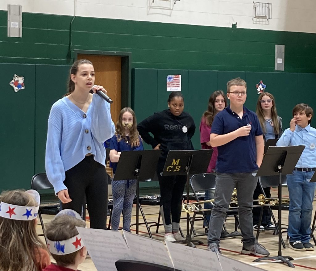 A student holds a microphone while others place their hands on their heart