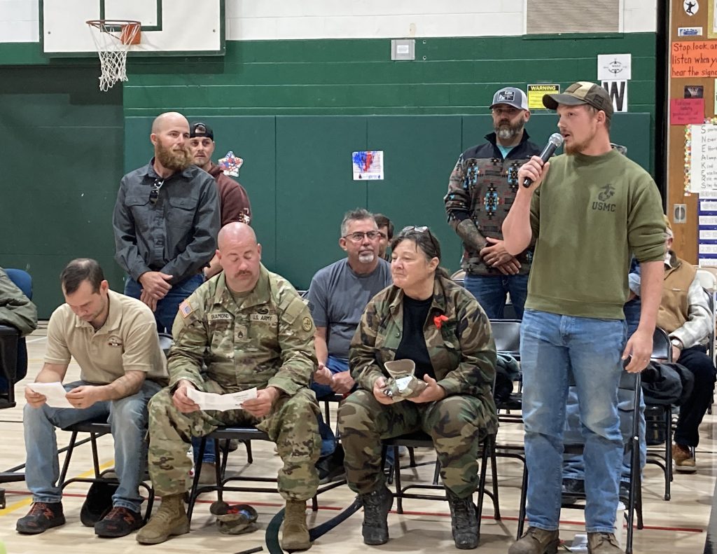 A veteran speaks into a microphone while other veterans, seated, look on