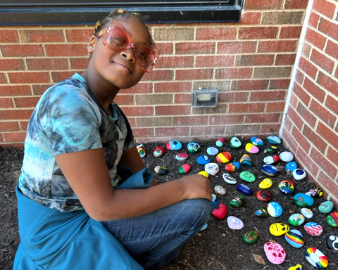 A girl with glasses and braids smiles as she kneels near a flowerbed filled with brightly painted rocks. 
