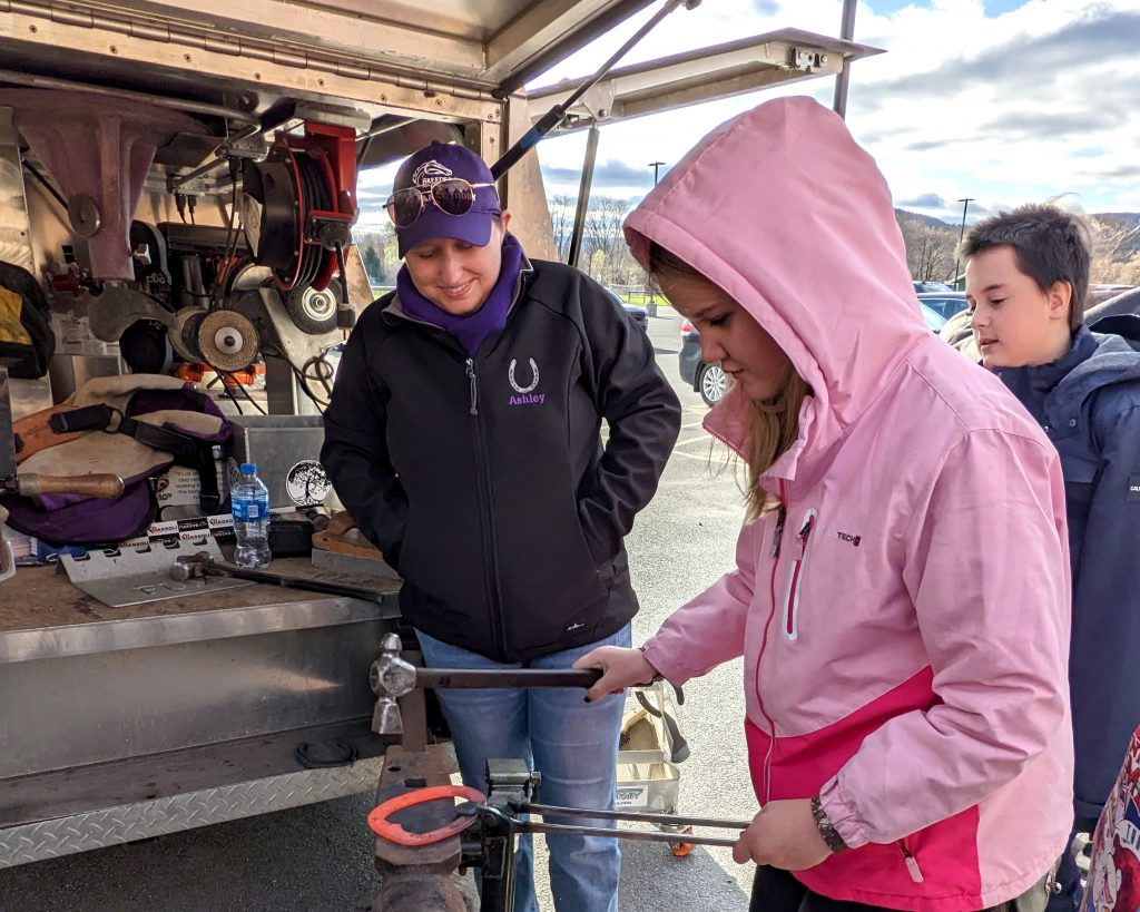 An adult looks on while a student tries her hand at hammering out a horseshoe