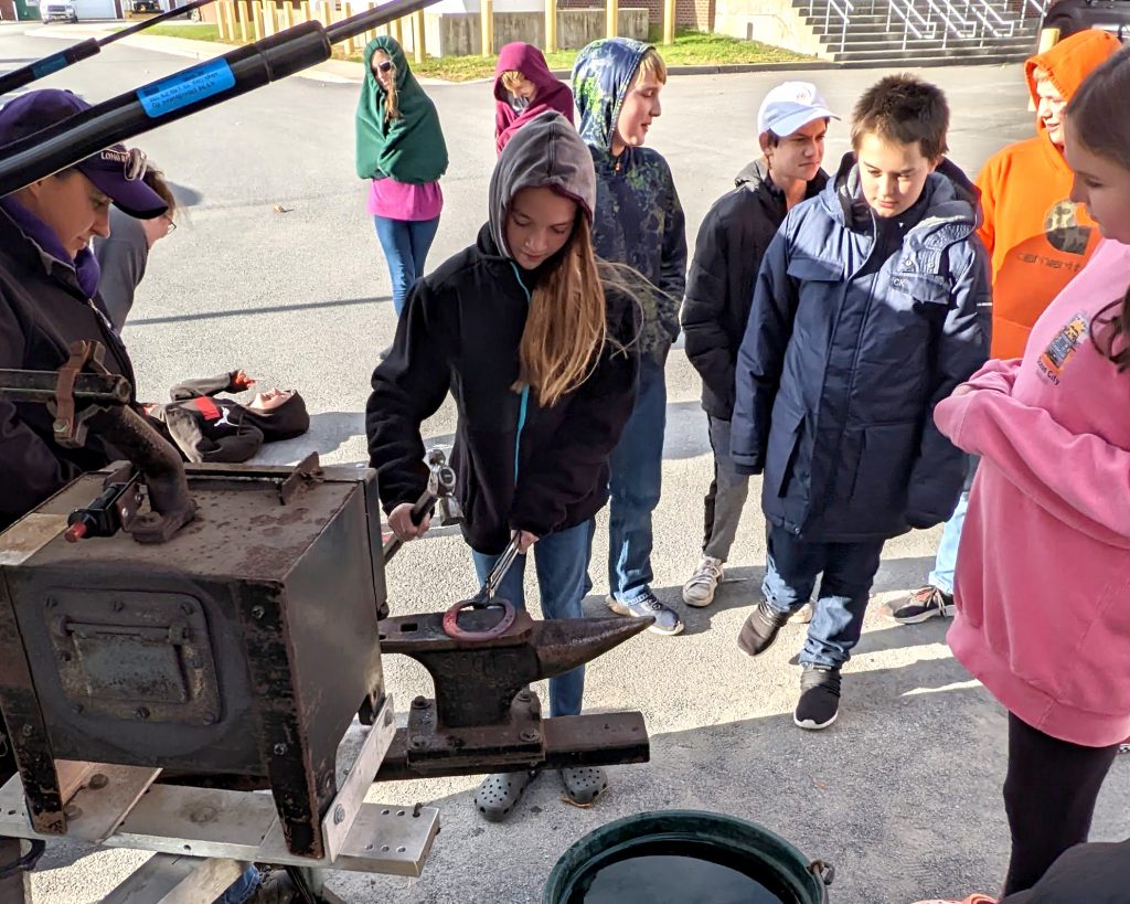 A student holds a red-hot horseshoe with tongs on a blacksmith's anvil while other students look on