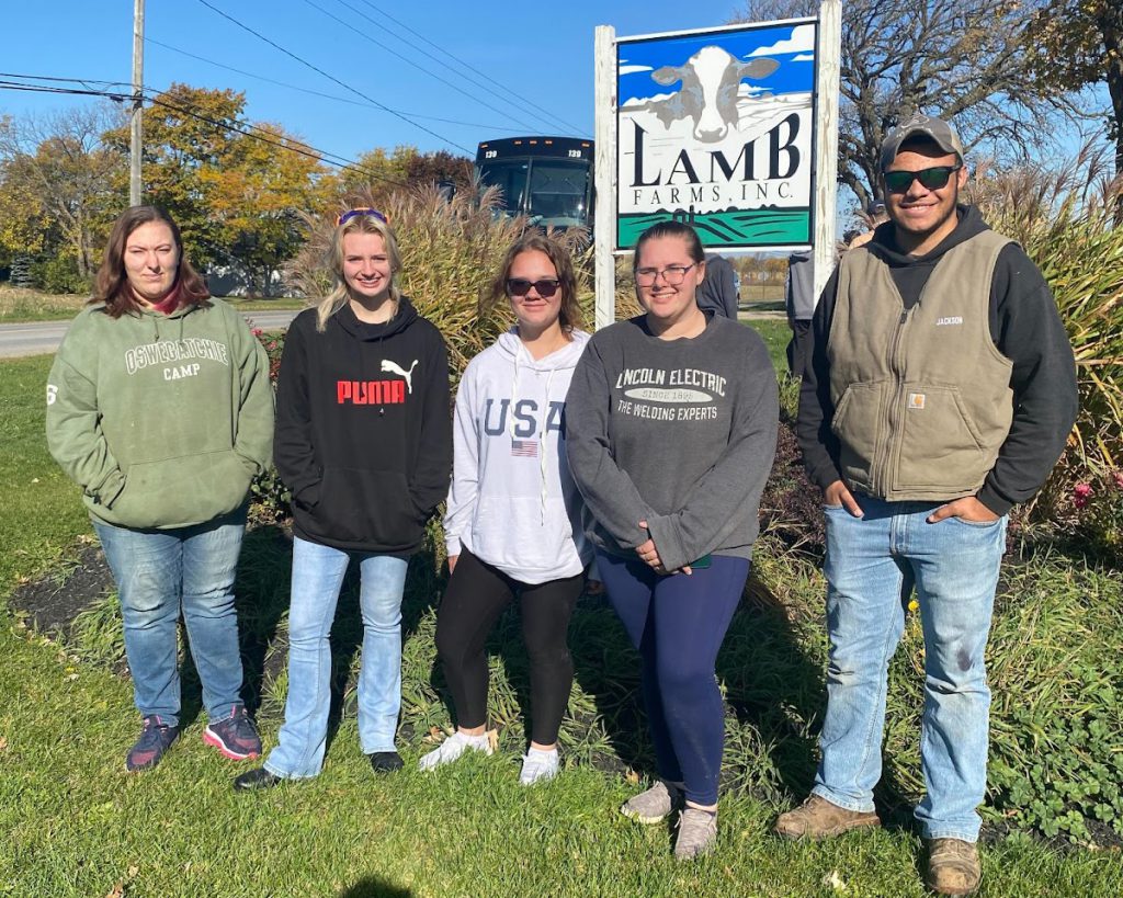 Students pose in front of a sign that reads Lamb Farms