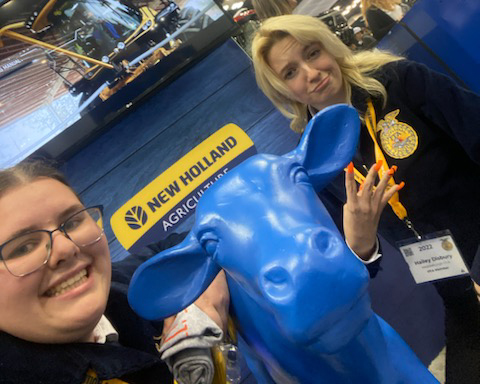 Two students wearing FFA jackets pose in front of a New Holland promotional display that includes a large blue cow