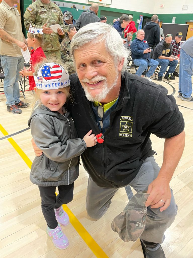 A man with white hair and beard smiles and kneels next to a little girl wearing a paper hat decorated with an American flag