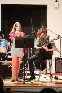 A teen girl sings next to a teen boy with a guitar.