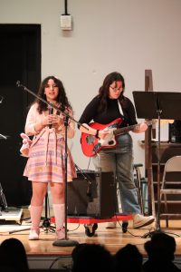Sisters stand on stage. One is singing, the other plays guitar.
