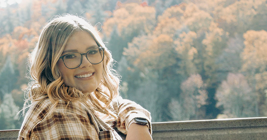 Teen girl smiles toward camera with autumn trees in the background.