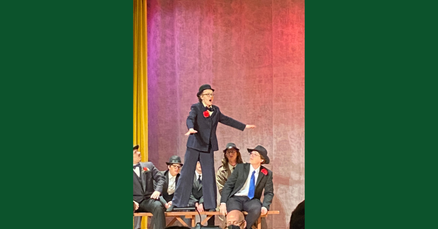 Student dressed in a suit, bowler het, and tie stands on a table with other students looking up at her during a theatrical performance.