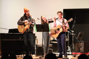 Two teens play guitar. Teen girl stands between them.