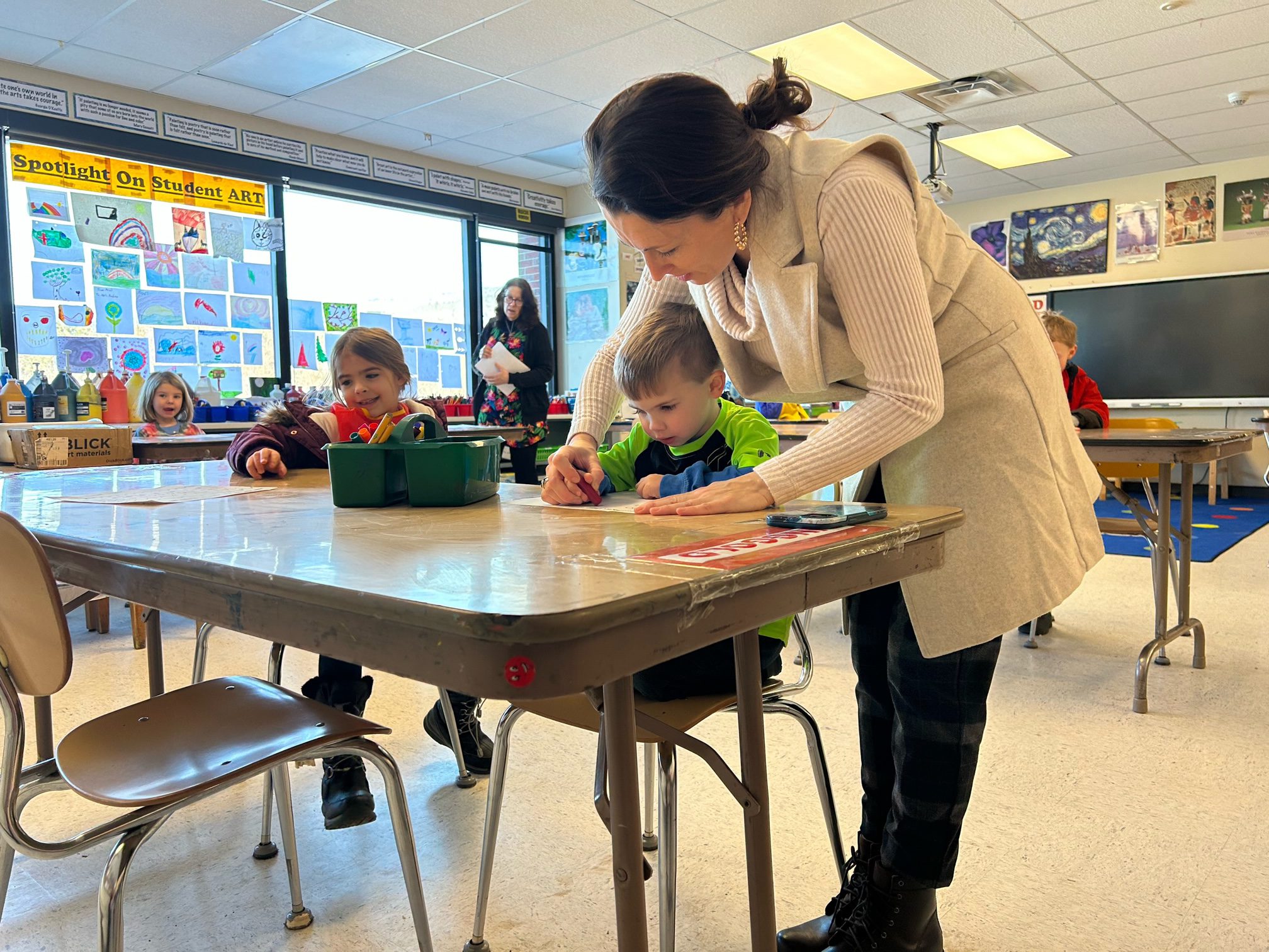 A teacher assists a student as another student looks on.