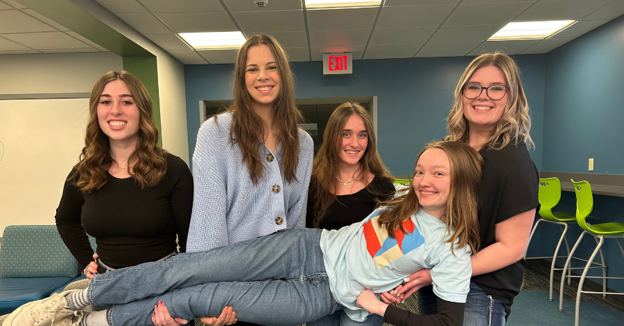 Four teenage girls hold another teenage girl up horizontally. They all smile.