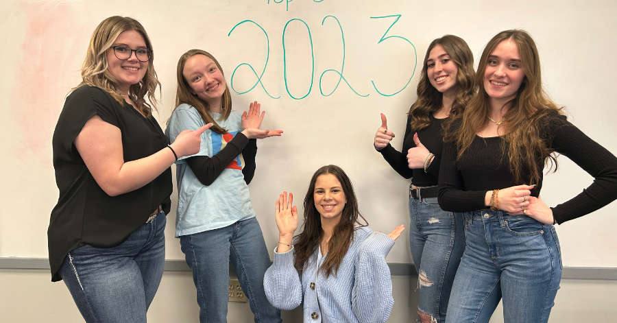 Five teenage girls stand around a dry erase board with the message 2023