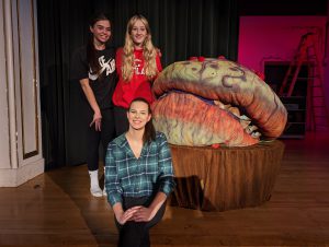 Three girls pose on stage with fake plant.