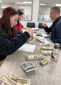 Studens work on a business assisngment. There is cash and a calculator on the desk. A teacher is present.