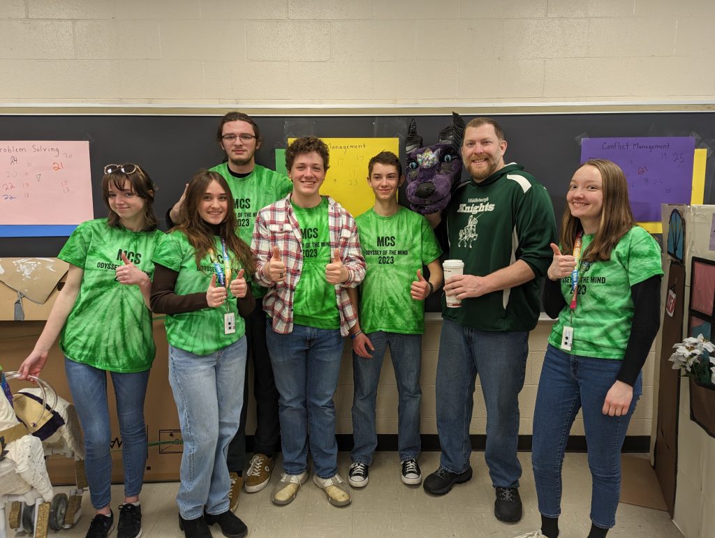 Teens give thumbs up. The group is wearing matching green t-shirts.