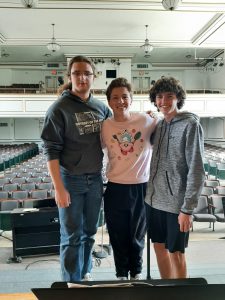 Three teens stand with arms around each other in front of auditorium seats.