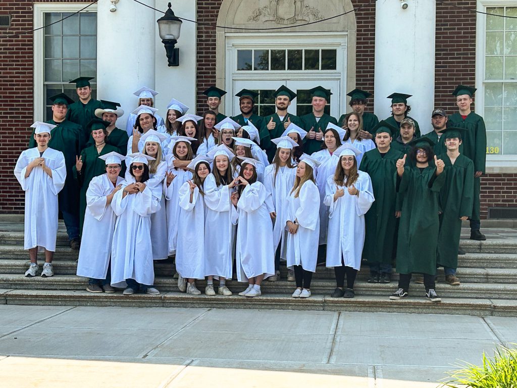 Class of 2023 assembled on steps of high school. They are wearing caps and gowns and making silly poses.