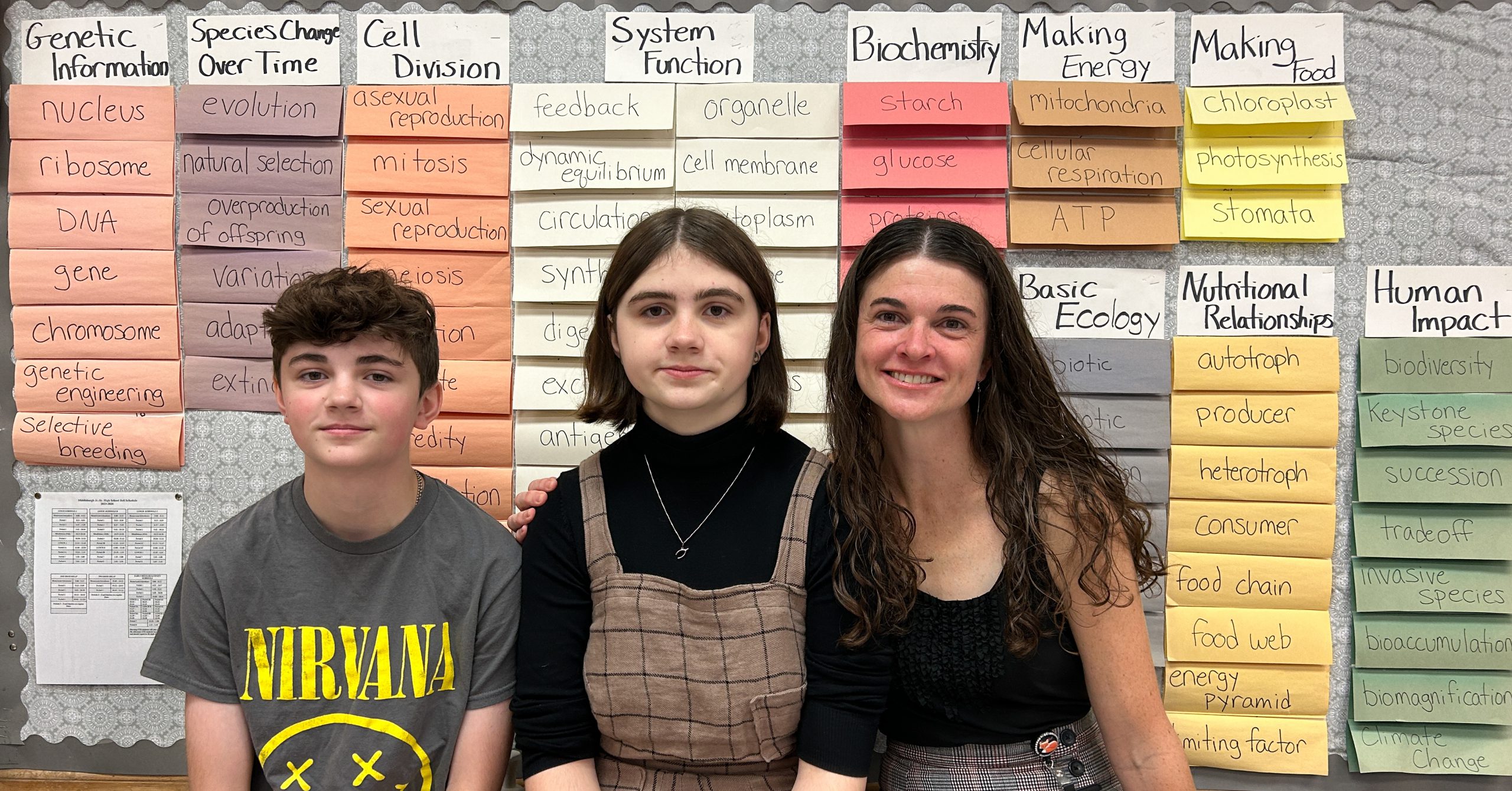 Teacher and two students in front of biology vocabulary bulletin board.
