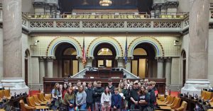 31 MCS students stand in the NYS Legislative session room.