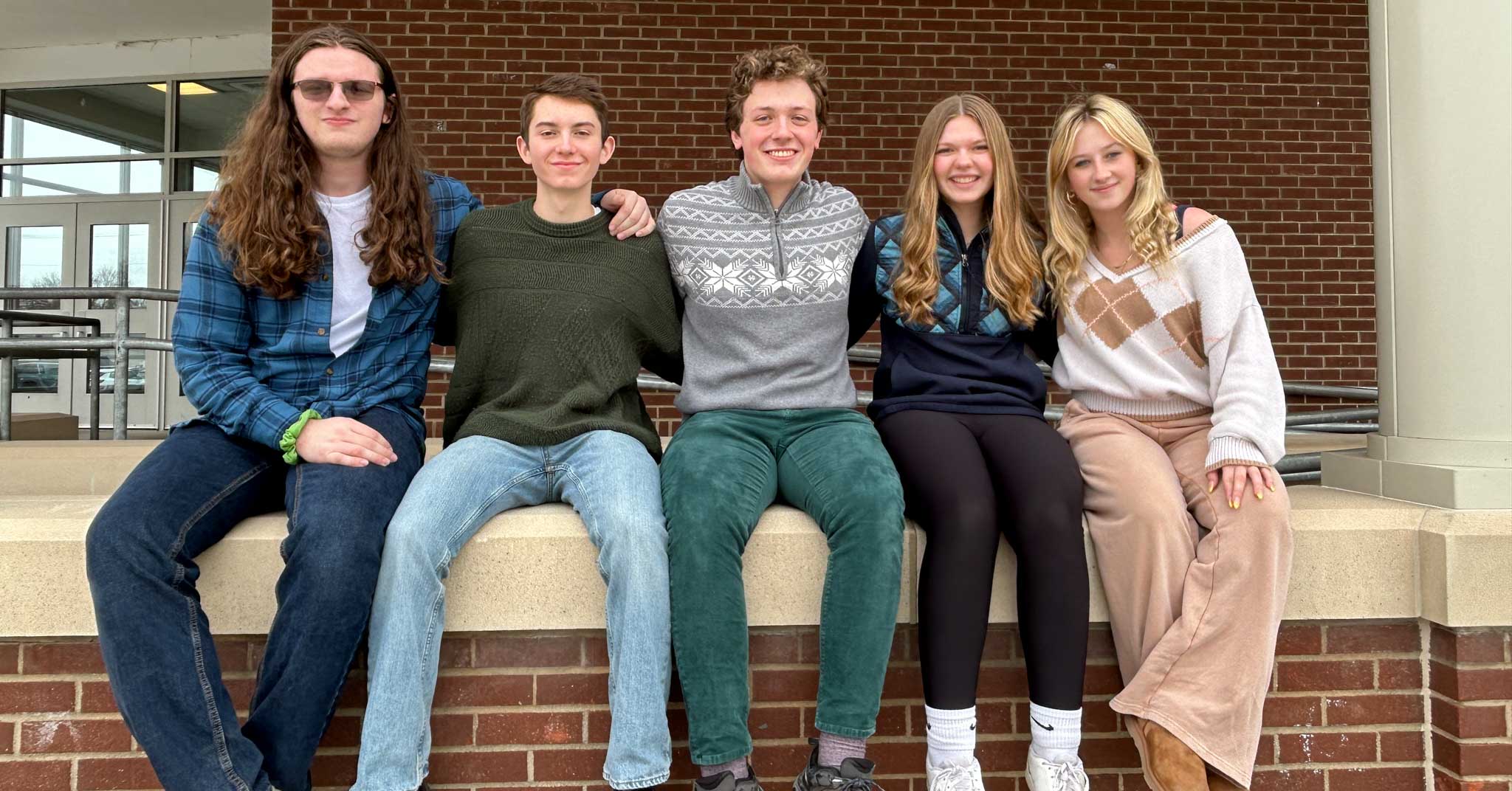 Five students sitting in a row on half row. They have arms around each other.