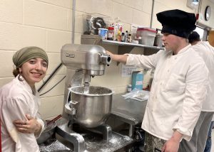 Two teens stand next to restaurant-sized food mixer.