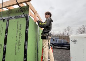 Student stands on ladder and works on frame of a tiny home.