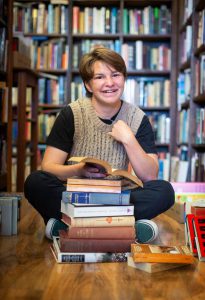 Student sits on floor of library with a stack of books in front of them.