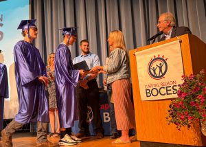 Students, dressed in cap and gowns, cross stage to receive award.