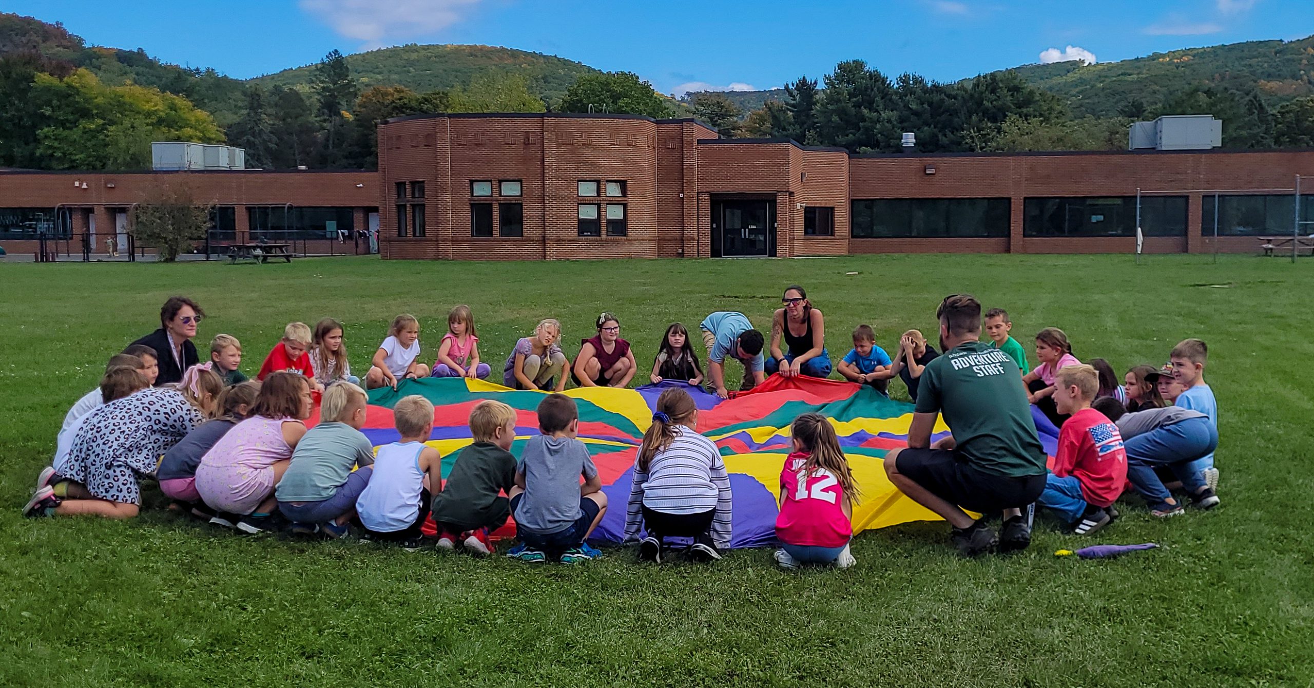 Elementary students surround a multi-colored parachute. School building in background.