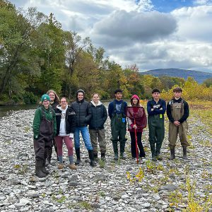 Group of teams, many wearing tall boots, stand on the shore of a river.