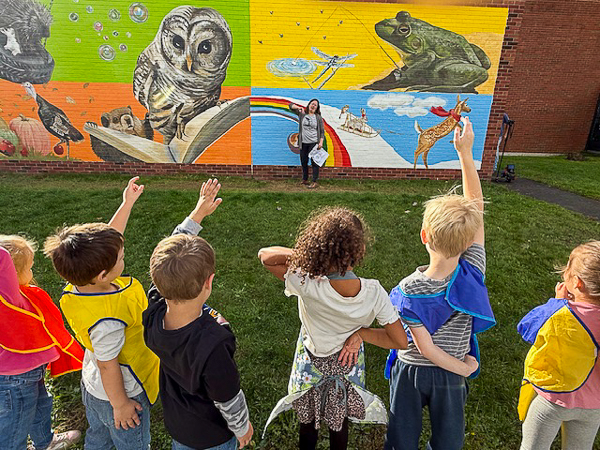 Artist speaks to children. Several children have their hands raised. Mural in background.
