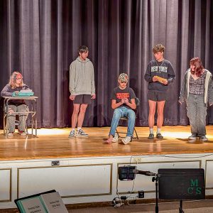 Two teens rehearse on stage, One sits behind a typewriter on a desk.