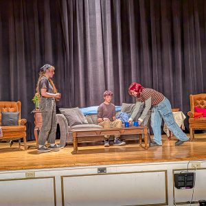 Three teens rehearse on staged living room set.