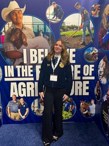 Student, wearing FFA jacket stands in front of "I believe in the future of agriculture" backfrop.