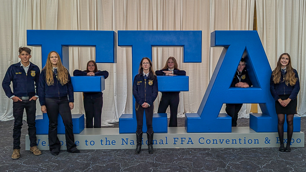 Students weraing FFA jackets stand in front of the FFA National Convention sign.