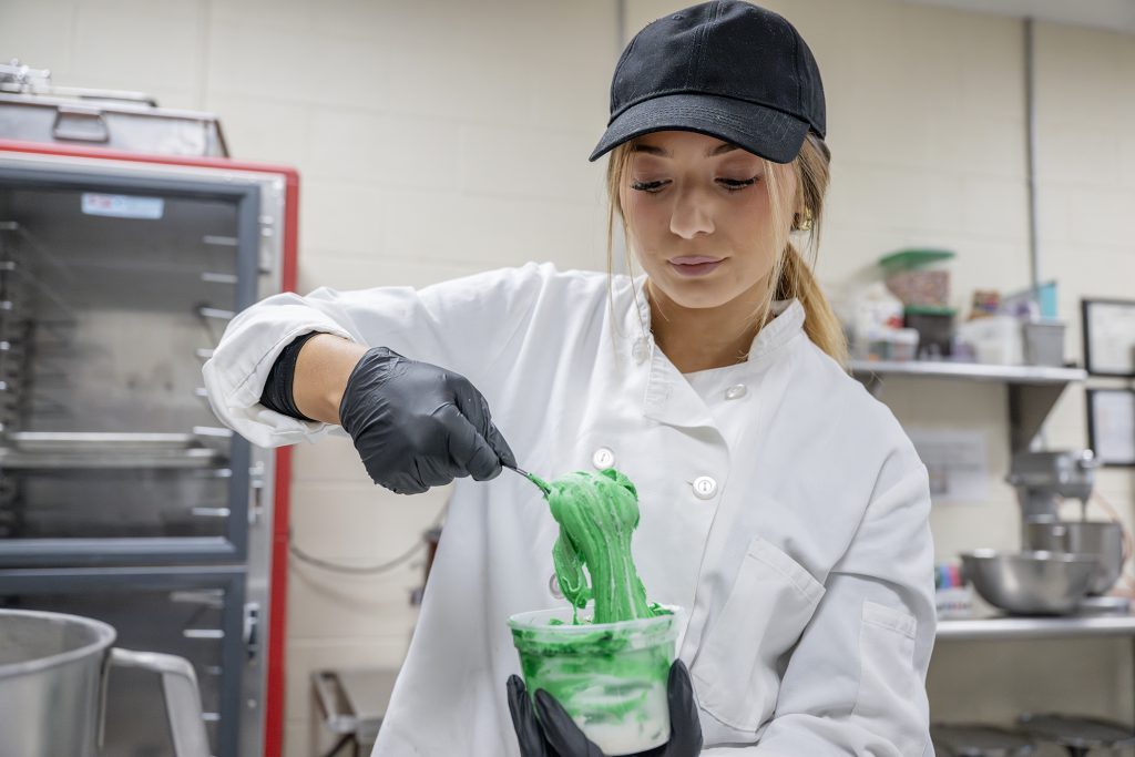 Student wearing gloves, a hat and a chef's jacket holds a container of green frosting.