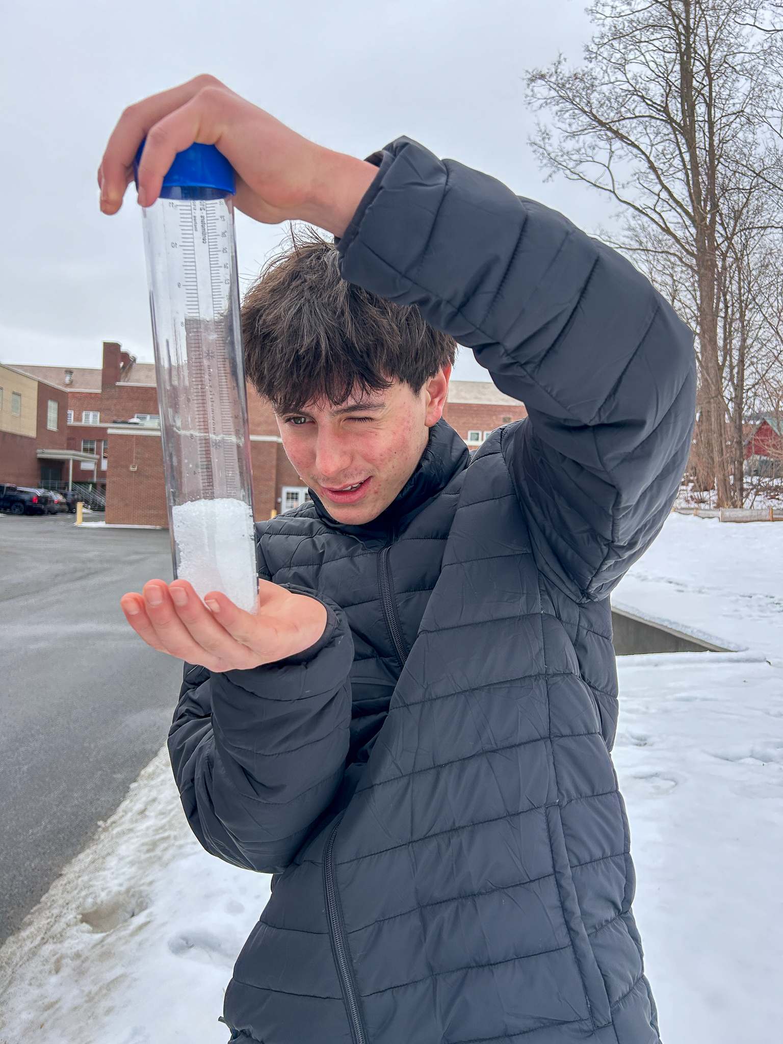 A student outdoors holds up a clear cylindrical tube partially filled with snow, examining the measurement markings on the side. Snow-covered ground and school buildings appear in the background.