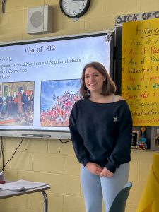 A student stands at the front of a classroom giving a presentation on the War of 1812. A large screen behind the student displays historical images and bullet points related to the conflict. Classroom posters and notes decorate the surrounding walls.