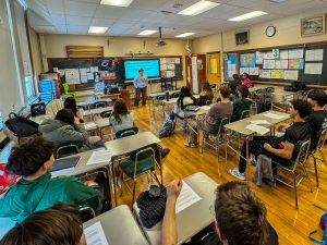 A classroom full of students sits at individual desks facing a teacher who is standing at the front of the room next to a projected slide. The room is decorated with student artwork, posters, and classroom materials, and the students appear to be engaged in a lesson.