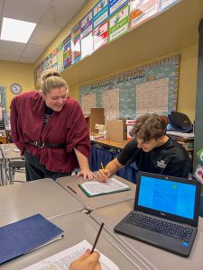 A teacher leans over a student’s desk to offer guidance as the student works. Another student’s hand is visible in the foreground holding a pencil. A laptop on the desk displays a map, and colorful instructional posters line the walls.