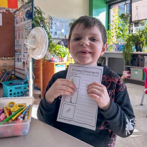 A child holds up a completed worksheet titled “Growing Ornaments” at a classroom desk. A container of markers, scissors, and other supplies is in the foreground, and plants are visible in the background.