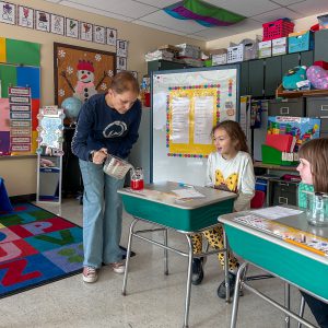 An adult pours red liquid from a measuring cup into a jar on a student’s desk. Two children sit at desks nearby, and the classroom background includes a whiteboard, colorful posters, and storage bins.