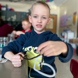 A child wearing a dark hoodie with a cartoon character design holds two pipe cleaners, one white and one brown, while sitting at a classroom desk. Other students and jars are visible in the background.