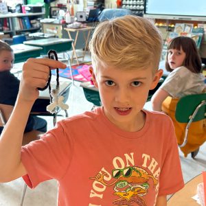 A child in a peach-colored shirt with a graphic design holds up a crystal formed on black and white pipe cleaners. The classroom setting includes desks, chairs, and colorful materials in the background.