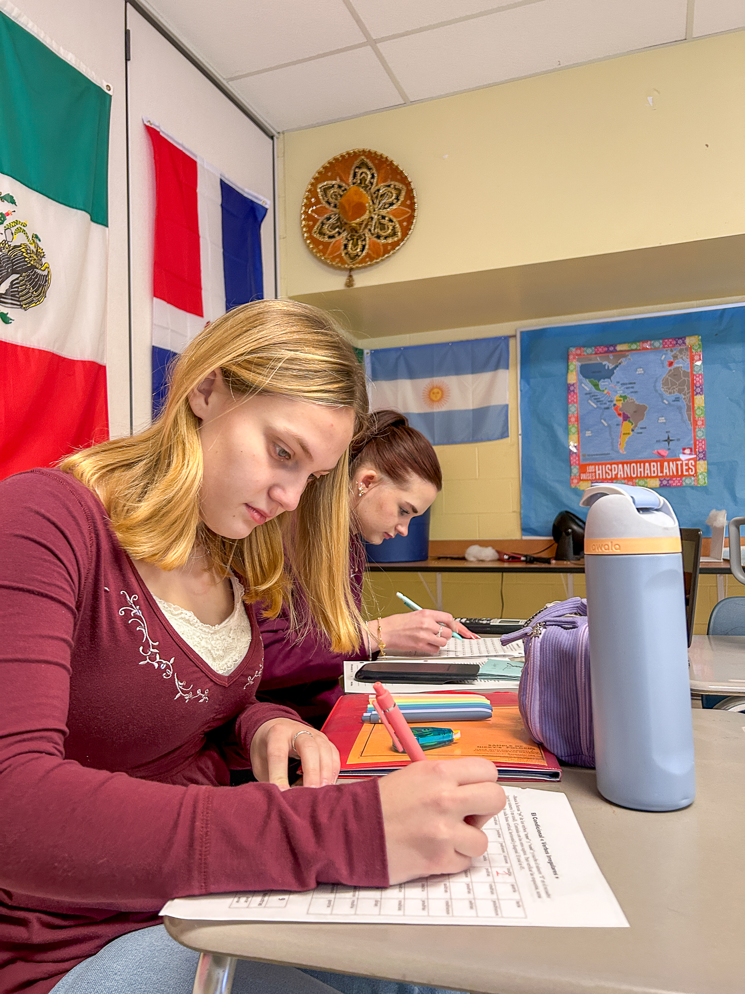 Two students sit at a classroom desk working on a Spanish assignment. One writes on a worksheet with a pink pen while textbooks, notebooks, and flags from Spanish‑speaking countries decorate the room.