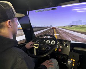 A student sits in a vehicle simulator, holding the steering wheel while a large screen displays a driving scenario on a multi‑lane road with the speed shown as 32 mph.