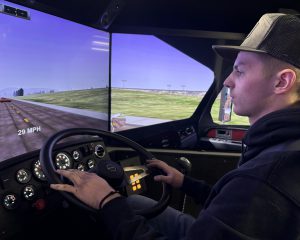 A student operates a vehicle simulator, gripping the steering wheel as a wide driving scene appears across curved display screens, showing the vehicle traveling at 29 mph.