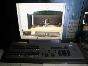 A view from a lighting booth featuring an ETC control console overlooking a theater stage with a green curtain. To the right, graffiti on the wall commemorates "Rick Coons & Jan Cooper" from 1992.